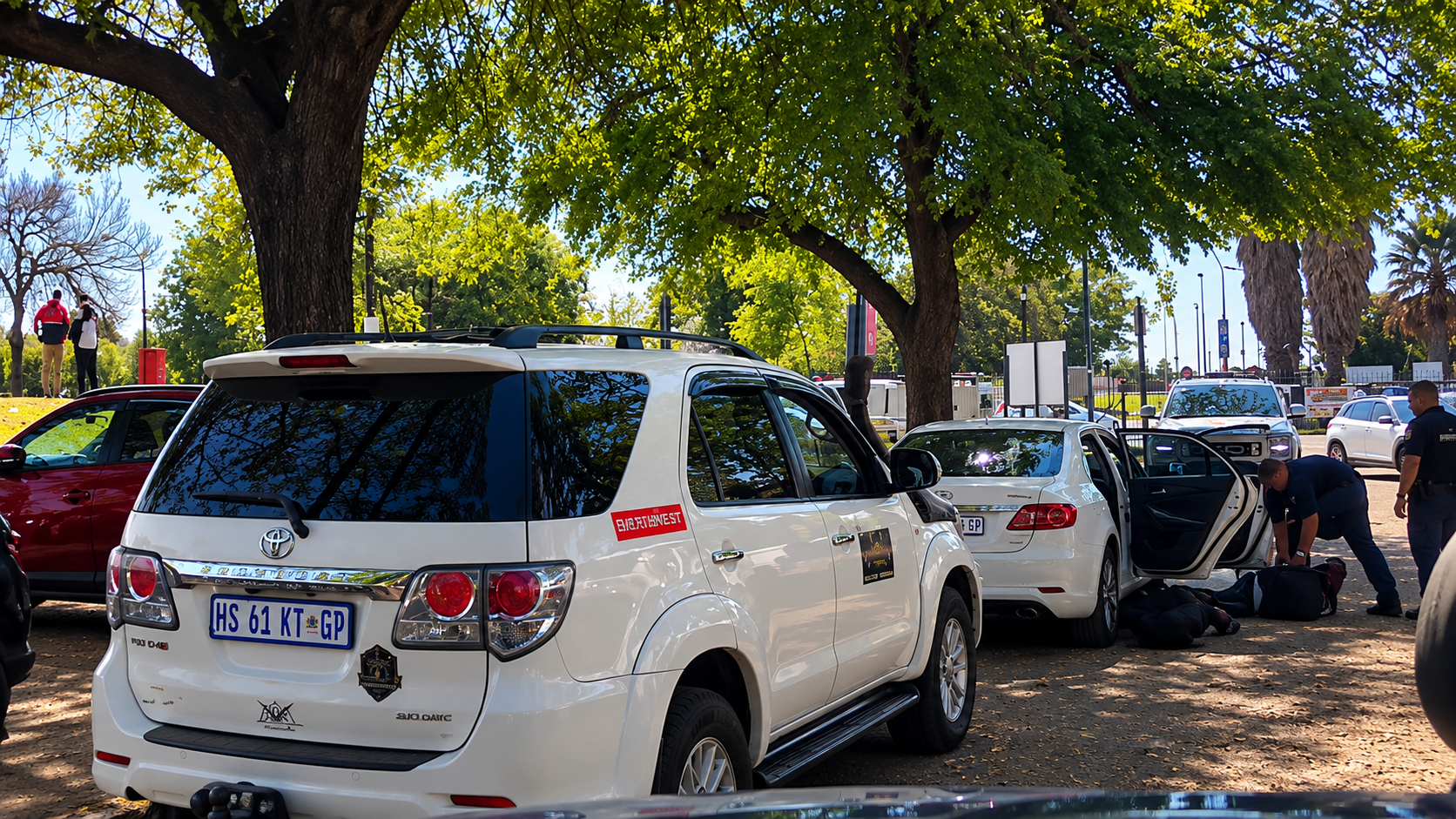 Members of the responding units with suspects on the ground beside a white sedan in a tree-lined parking area