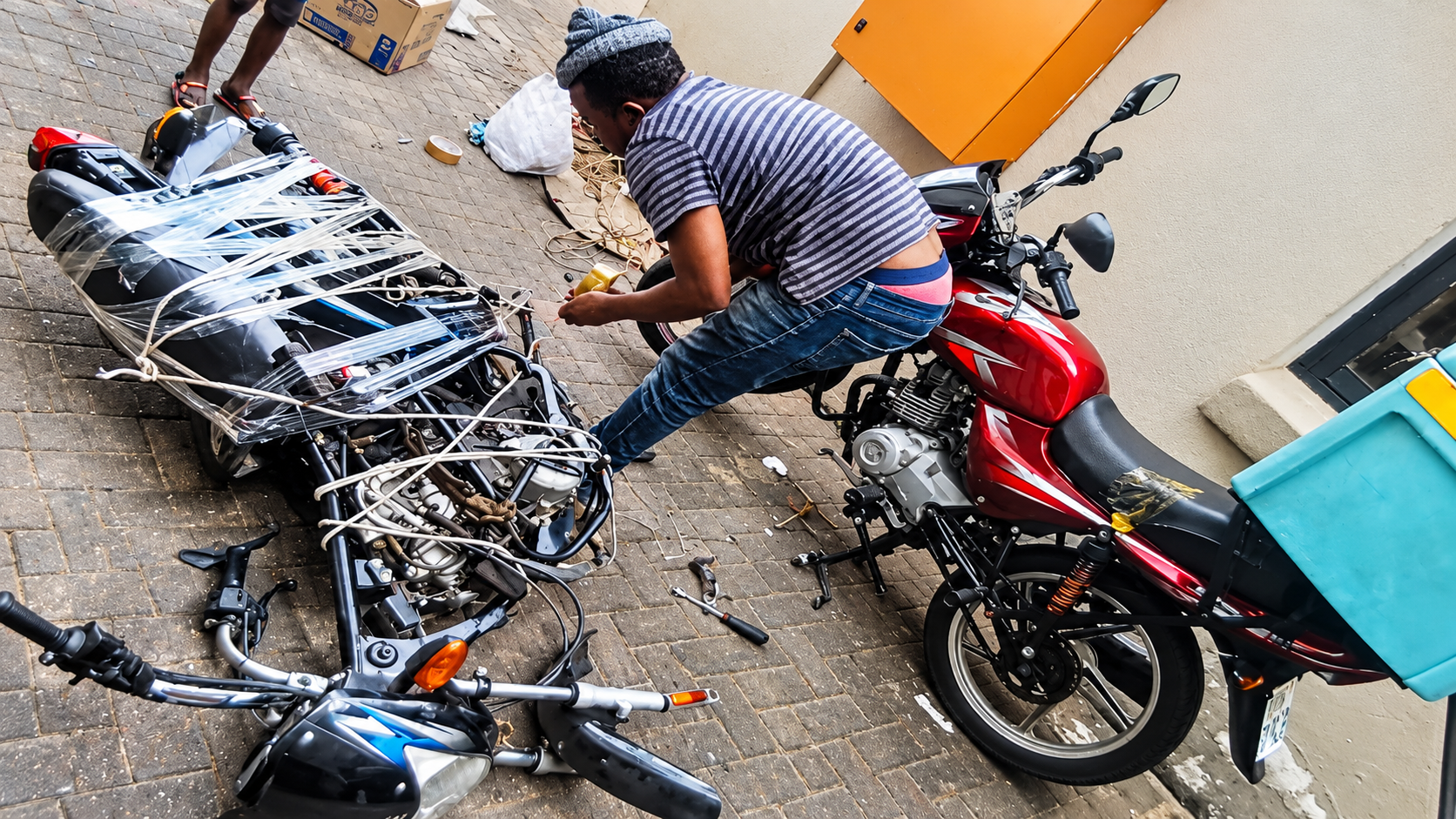 A motorcycle stripped down to its frame and engine, wrapped in plastic and tied with rope on a paved yard